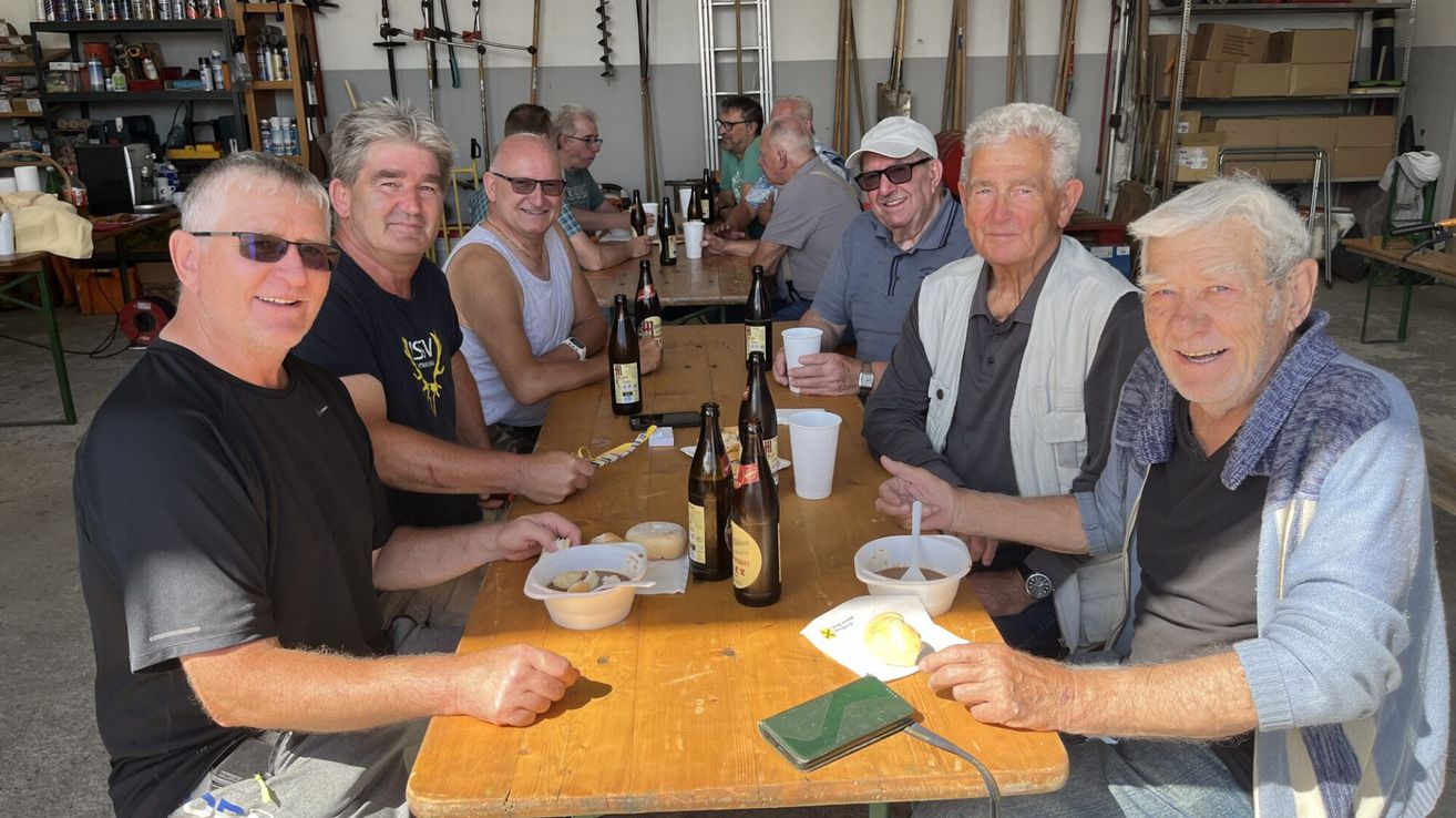 Eine Gruppe älterer Männer versammelt sich um einen Tisch, lächelt und posiert für ein Foto. Sie genießen ihr Essen und Getränke. Der Tisch ist mit Flaschen, Bechern und Tellern gedeckt. Hinter ihnen stehen verschiedene Werkzeuge und Ausrüstung.