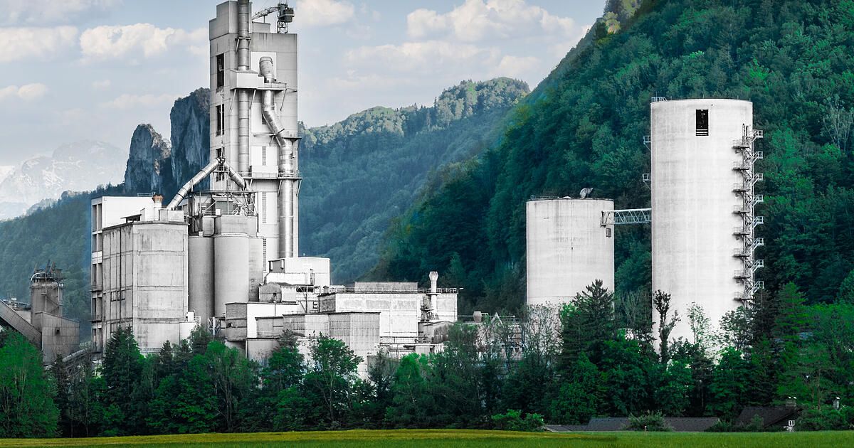 Ein großes Industriegebäude mit mehreren hohen Strukturen ist von einem üppigen Wald und Bergen unter einem blauen Himmel umgeben.