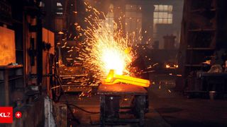 Ein Arbeiter in einer Fabrik formt mit einem Schweißbrenner ein glühendes Metallstück auf einem Tisch, wobei Funken aufsteigen.