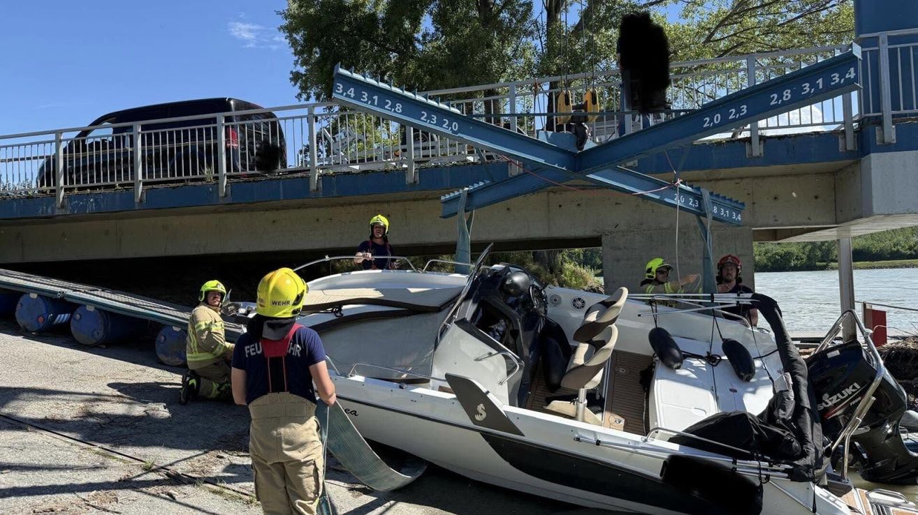 Ein beschädigtes Boot wird mit Seilen angehoben. Feuerwehrleute mit gelben Helmen sind am Unfallort. Ein Auto befindet sich auf einer Brücke über dem Boot.