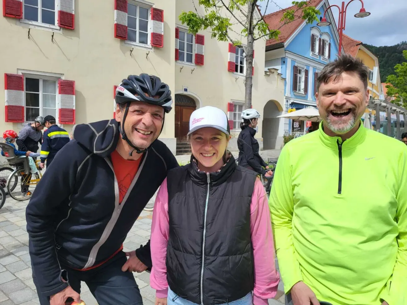 Drei Personen stehen vor einem Gebäude mit roten Fensterläden und lächeln. Einer trägt einen Helm, ein anderer eine Kappe und der Dritte ein neongrünes Jacket. Eine Person fährt hinter ihnen auf einem Fahrrad.
