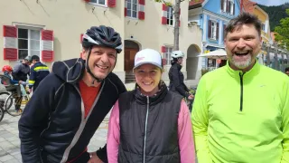 Drei Personen stehen vor einem Gebäude mit roten Fensterläden und lächeln. Einer trägt einen Helm, ein anderer eine Kappe und der Dritte ein neongrünes Jacket. Eine Person fährt hinter ihnen auf einem Fahrrad.