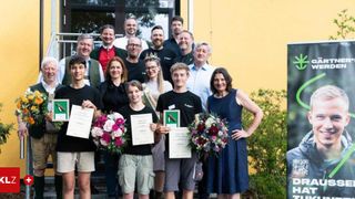 Eine Gruppe von Menschen steht vor einem Gebäude mit Blumen und Urkunden. Sie lächeln und halten Urkunden in den Händen. Sie feiern wahrscheinlich eine Leistung.