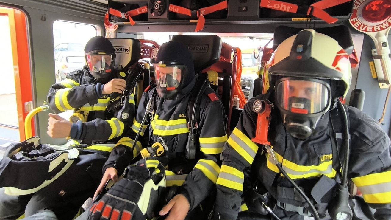 Drei Feuerwehrleute in voller Ausrüstung sitzen in einem Fahrzeug. Jeder trägt Helm, Maske und Handschuhe. Ausrüstung und Werkzeuge sind um sie herum sichtbar.