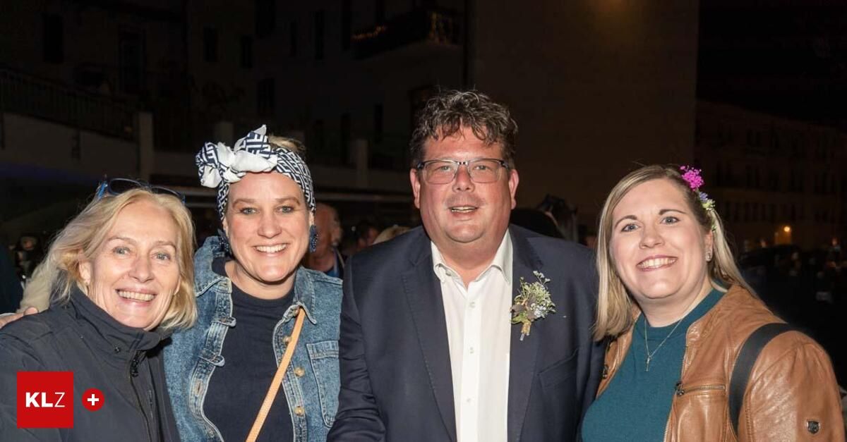 Eine Gruppe von Menschen steht zusammen und lächelt für ein Foto. Ein Mann trägt einen Anzug und eine Ansteckblume, während zwei Frauen Jeansjacken tragen. Der Hintergrund ist verschwommen mit Leuten.