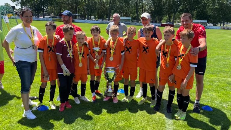 Eine Gruppe junger Fußballspieler in orangefarbenen Trikots posiert mit ihren Medaillen und Pokalen auf einem Rasenfeld. Zwei Erwachsene, einer mit Kappe und der andere mit Hut, stehen hinter ihnen.