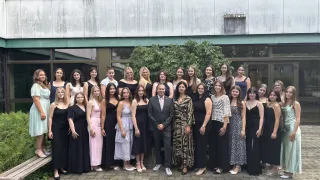 Eine Gruppe von Frauen und einem Mann, die formell gekleidet sind, stehen vor einem Gebäude mit großen Fenstern. Sie scheinen für ein Gruppenfoto zu posieren.