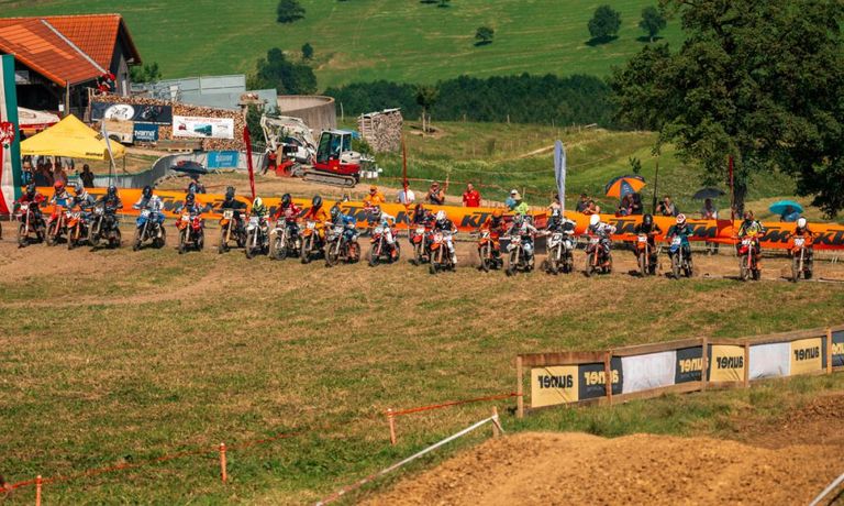 Ein Motorradrennen auf einer Dirt-Track-Rennstrecke, bei dem die Fahrer in Helmen bereit zum Start stehen. Zuschauer stehen hinter Barrieren. Im Hintergrund befinden sich Bäume, ein Gebäude und Maschinen.