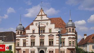 Ein altes Gebäude mit rotem Dach und weißen Wänden, mit großen Fenstern, kunstvollen Verzierungen und einer Uhr in der Mitte. Das Gebäude hat drei kuppelförmige Türme an der Vorderseite.
