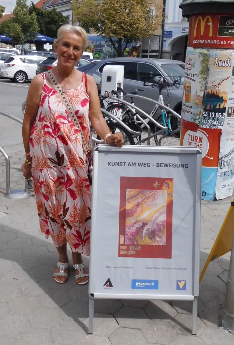 Eine Frau in einem Blumenkleid steht neben einem Plakat mit dem Titel Kunst am Weg. Das Plakat zeigt ein Gemälde und den Text Kunst am Weg - Bewegung. Dahinter stehen geparkte Fahrräder und eine Metallbarriere.