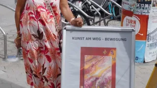 Eine Frau in einem Blumenkleid steht neben einem Plakat mit dem Titel Kunst am Weg. Das Plakat zeigt ein Gemälde und den Text Kunst am Weg - Bewegung. Dahinter stehen geparkte Fahrräder und eine Metallbarriere.