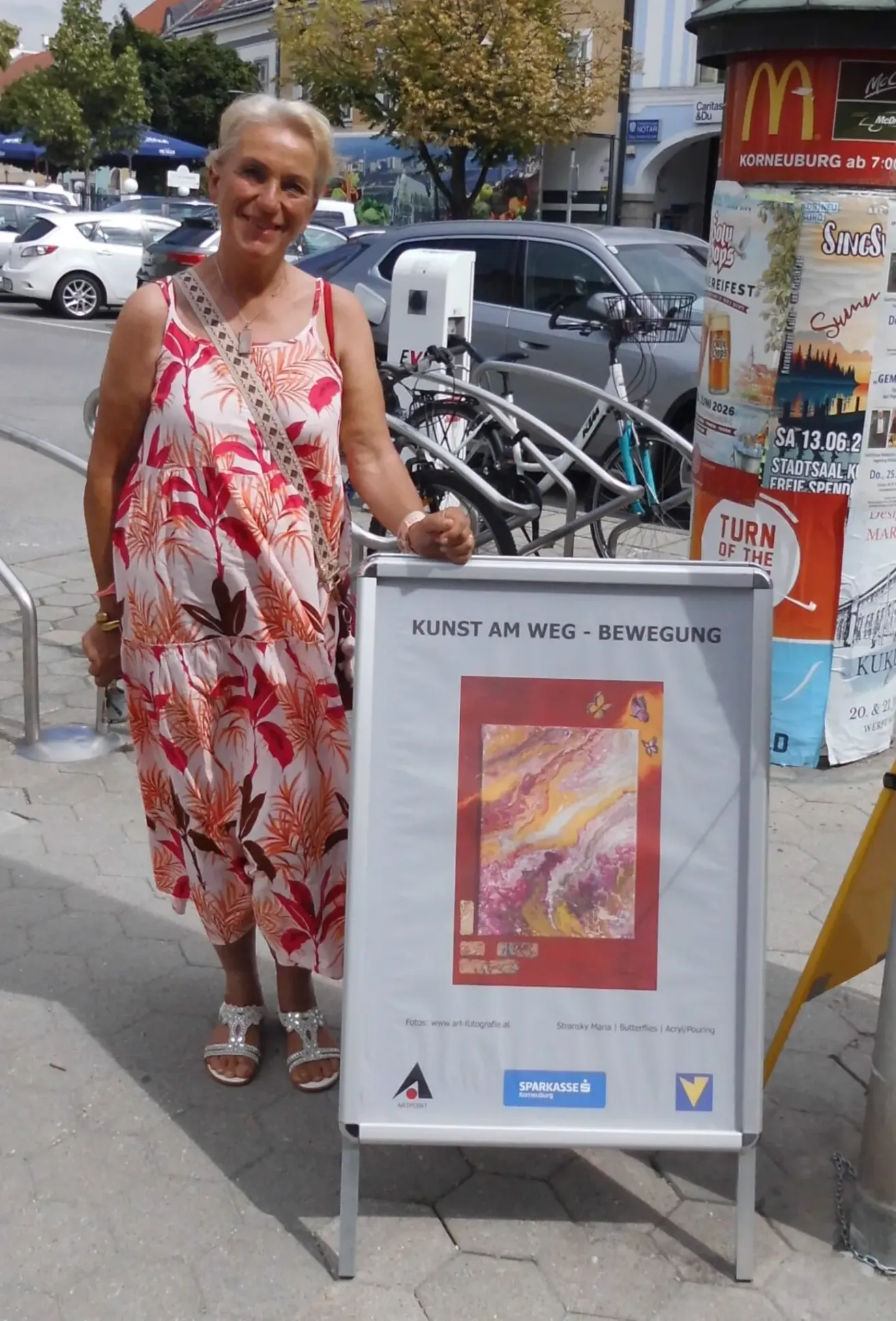 Eine Frau in einem Blumenkleid steht neben einem Plakat mit dem Titel Kunst am Weg. Das Plakat zeigt ein Gemälde und den Text Kunst am Weg - Bewegung. Dahinter stehen geparkte Fahrräder und eine Metallbarriere.