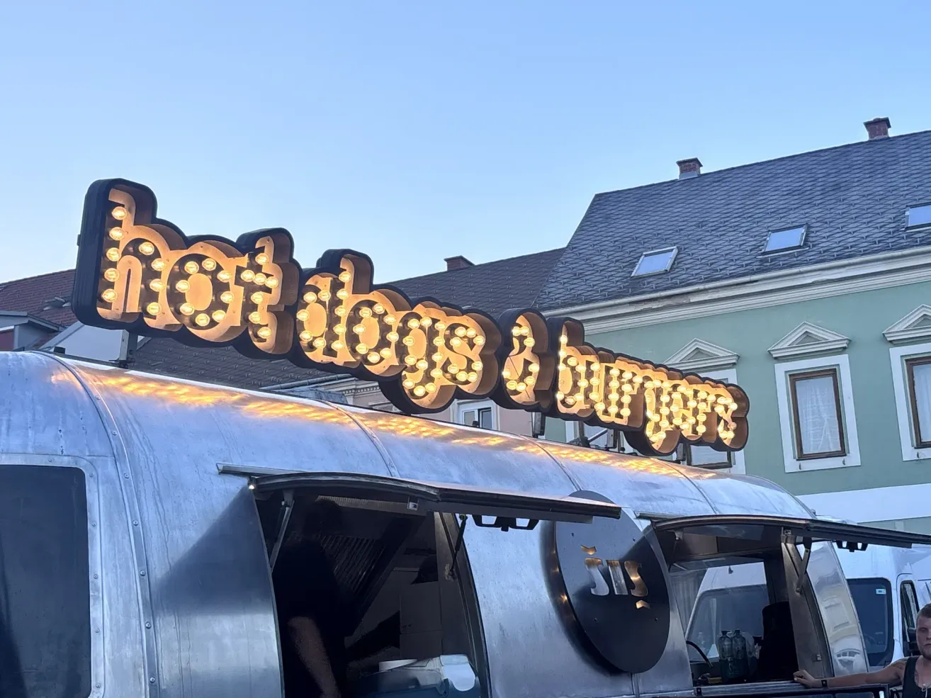 Ein Imbisswagen mit einem beleuchteten Schild, das 'Hot Dogs & Burger' anzeigt. Es hat eine offene Tür mit einer Person im Inneren. Der Wagen steht vor einem Gebäude mit Fenstern und einem Schornstein.