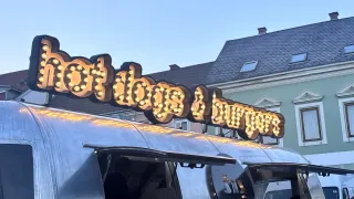 Ein Imbisswagen mit einem beleuchteten Schild, das 'Hot Dogs & Burger' anzeigt. Es hat eine offene Tür mit einer Person im Inneren. Der Wagen steht vor einem Gebäude mit Fenstern und einem Schornstein.