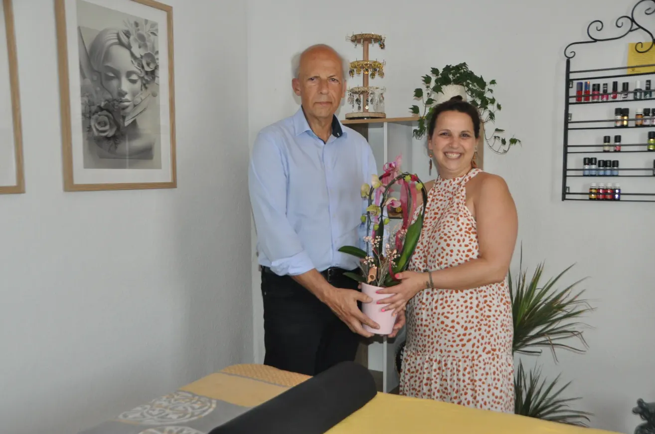 Ein Mann und eine Frau stehen in einem Raum und lächeln. Der Mann hält einen rosa Blumentopf. Sie stehen neben einer weißen Wand mit einem Bilderrahmen und einer Pflanze.
