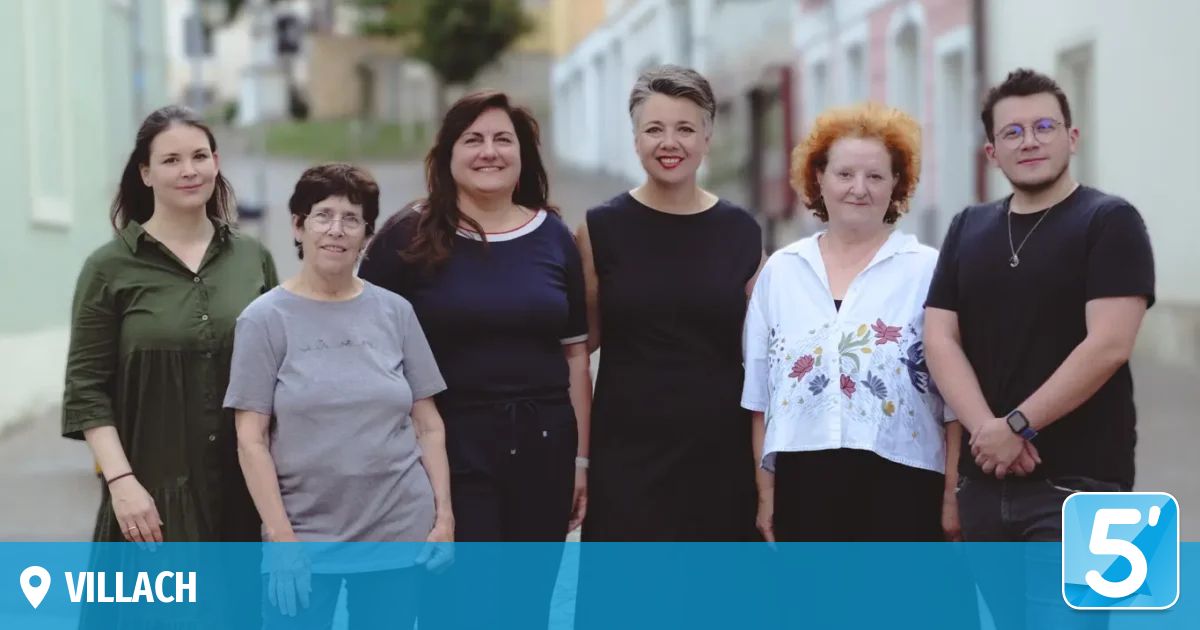 Fünf Frauen stehen zusammen auf einer Straße und lächeln für ein Foto. Hinter ihnen befinden sich Gebäude und Grünflächen.