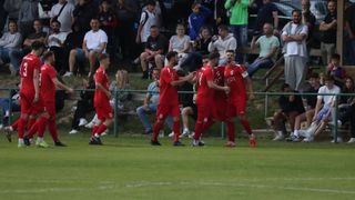 Eine Gruppe von Fußballspielern in roten Trikots feiert ein Tor vor einem Publikum, das auf Bänken sitzt. Die Spieler stehen auf dem Feld, während das Publikum zusieht.