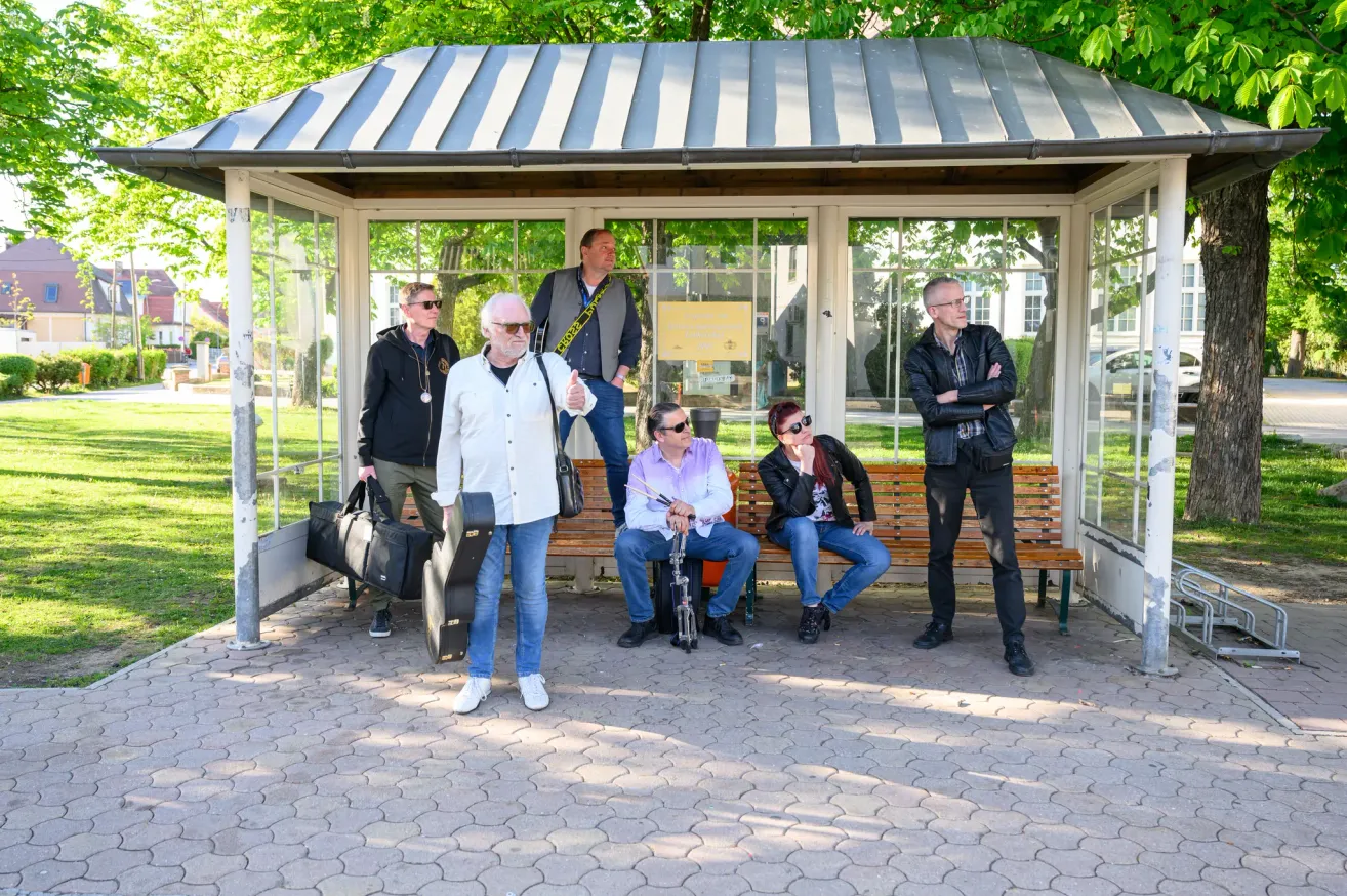 Fünf Personen versammeln sich in einem überdachten Bereich. Zwei sitzen auf einer Bank, während andere stehen, einer hält eine Gitarre. Ein Mann in einem weißen Mantel und einer Brille steht neben einem Mann mit einem Gitarrenkoffer.