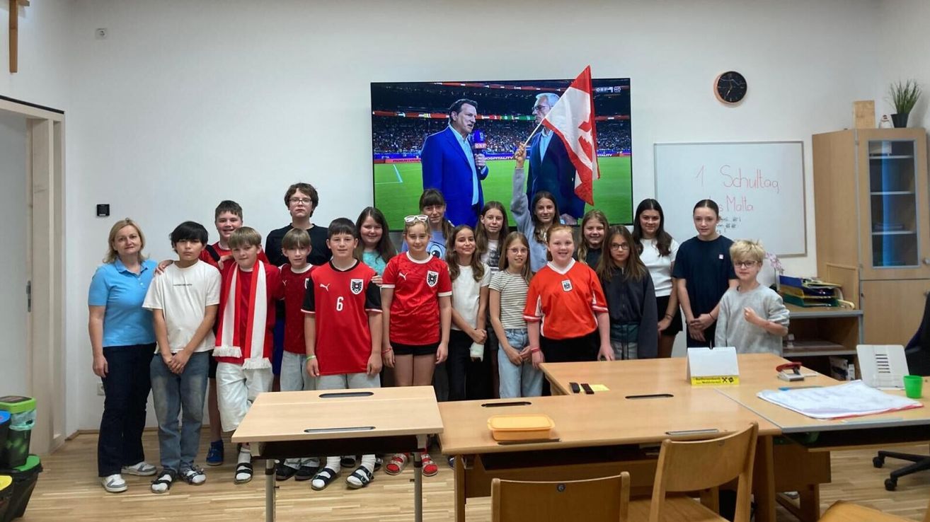 Eine Gruppe von Kindern in Sporttrikots posiert für ein Foto in einem Klassenzimmer mit einem großen Fernsehbildschirm hinter ihnen, der eine Sportveranstaltung zeigt.