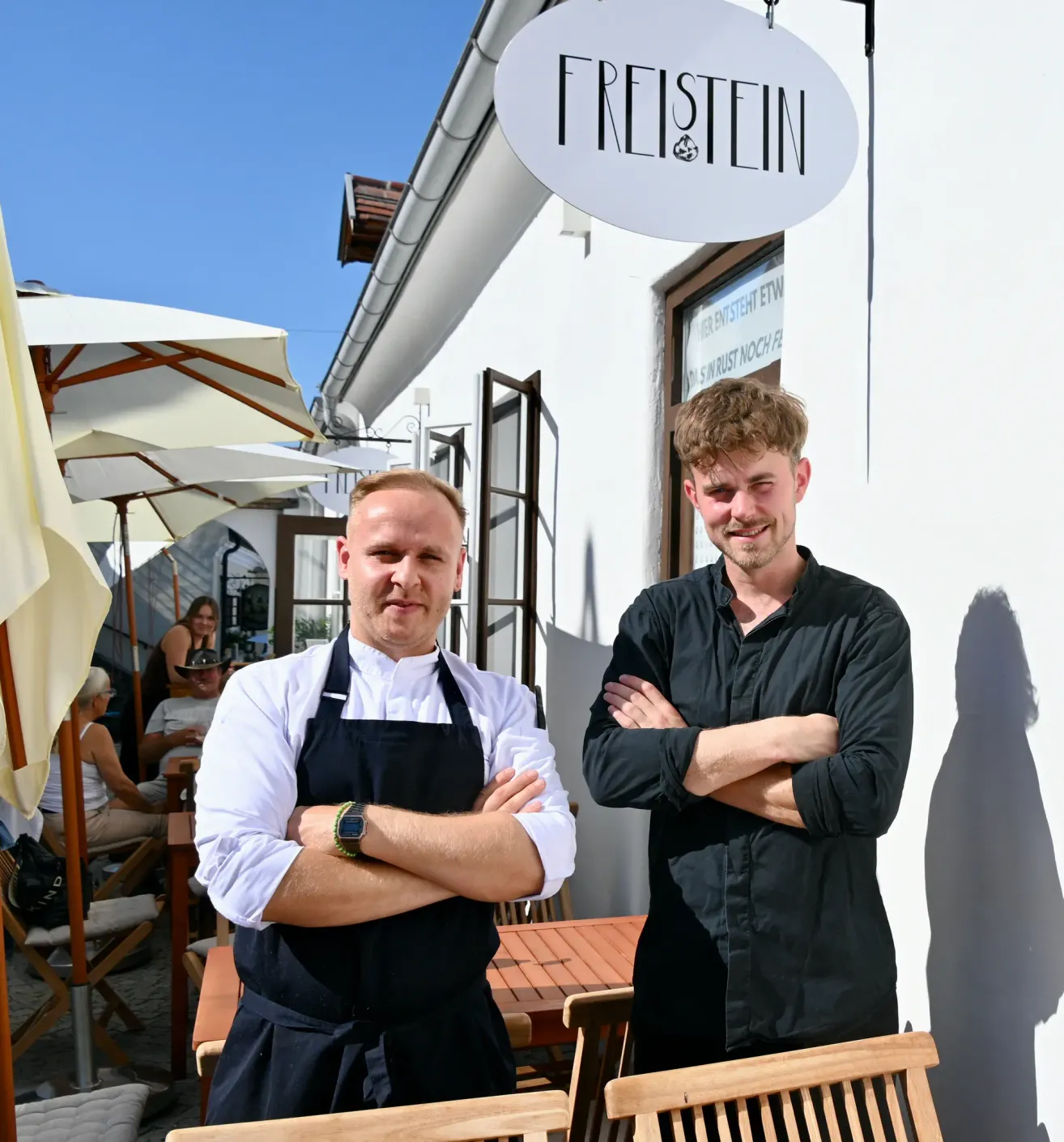 Zwei Männer stehen vor einem Gebäude mit dem Namen Freistein. Sie lächeln und einer hat die Arme verschränkt. Hinter ihnen sind Schirme und Tische mit sitzenden Personen.