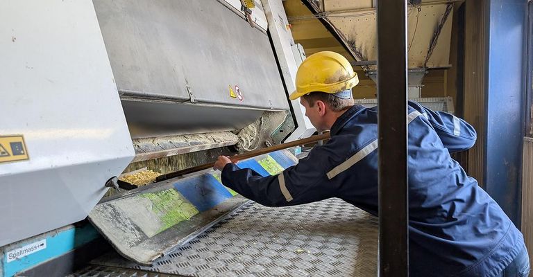 Ein Arbeiter in einer Fabrik mit einem gelben Helm benutzt einen langen Stab, um eine Substanz in einen Metallbehälter zu leiten.
