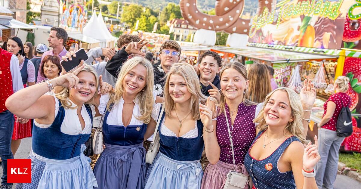 Eine Gruppe junger Frauen und Männer in traditioneller bayerischer Kleidung, die bei einem Festival lächelnd und posierend für ein Foto, mit bunten Fahrgeschäften im Hintergrund.