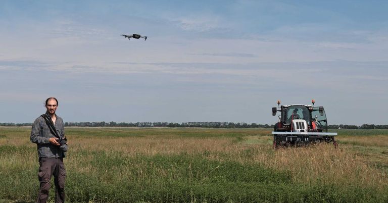 Ein Drohnen fliegt über einem Traktor, der in einem weiten Feld unter einem blauen Himmel mit Wolken Pflanzen ernte.