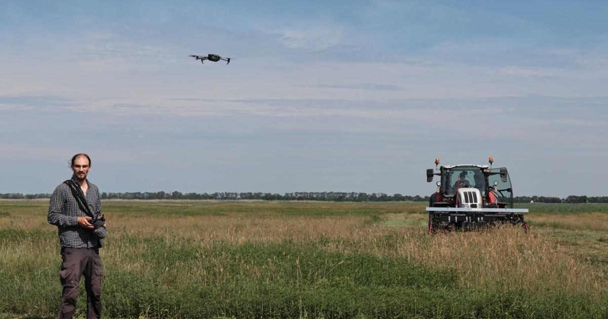 Ein Drohnen fliegt über einem Traktor, der in einem weiten Feld unter einem blauen Himmel mit Wolken Pflanzen ernte.