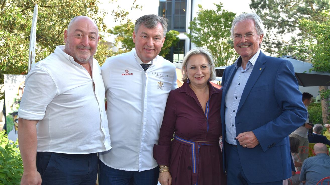 Vier Personen lächeln für ein Foto in einer Gartenumgebung. Der Mann im weißen Kochmantel trägt ein Michelin-Logo. Dahinter steht ein Gebäude mit Glasfenstern.