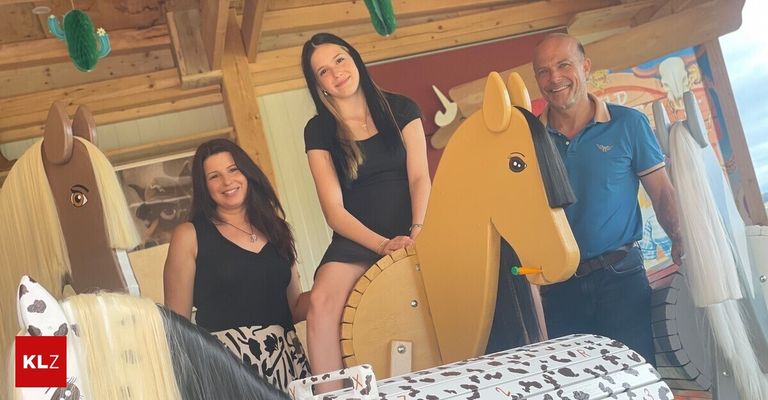 Drei Menschen posieren für ein Foto in einem hölzernen Raum, eine Frau sitzt auf einem Spielzeugpferd, eine andere Frau steht neben ihr, und ein Mann steht rechts.