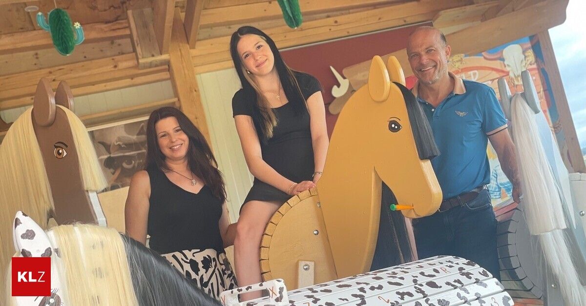 Drei Menschen posieren für ein Foto in einem hölzernen Raum, eine Frau sitzt auf einem Spielzeugpferd, eine andere Frau steht neben ihr, und ein Mann steht rechts.