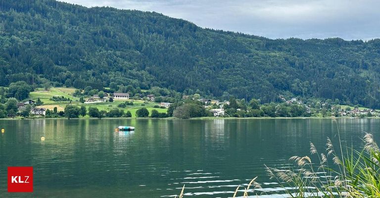 Ein ruhiger See umgeben von üppigen grünen Hügeln und Wäldern, mit einigen Gebäuden in der Ferne unter einem bewölkten Himmel. Ein kleines aufblasbares Floß schwimmt auf dem Wasser.