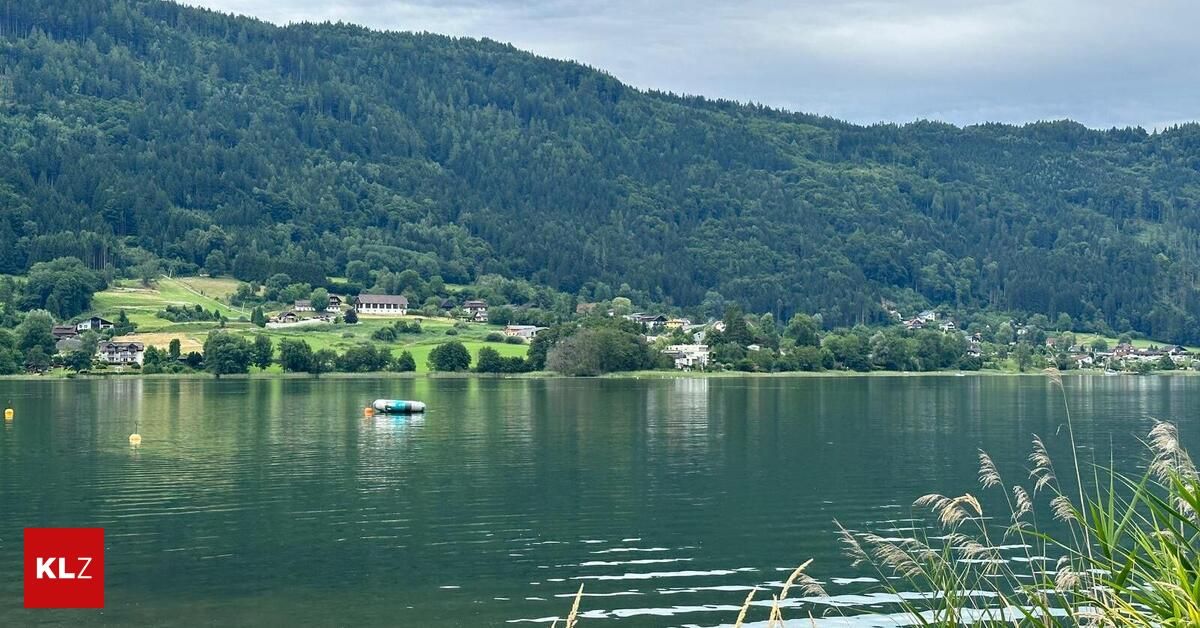 Ein ruhiger See umgeben von üppigen grünen Hügeln und Wäldern, mit einigen Gebäuden in der Ferne unter einem bewölkten Himmel. Ein kleines aufblasbares Floß schwimmt auf dem Wasser.