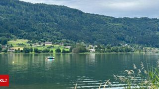 Ein ruhiger See umgeben von üppigen grünen Hügeln und Wäldern, mit einigen Gebäuden in der Ferne unter einem bewölkten Himmel. Ein kleines aufblasbares Floß schwimmt auf dem Wasser.
