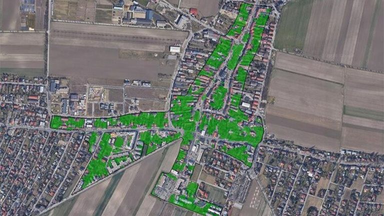 Luftaufnahme einer Stadt mit vielen Gebäuden und Grünflächen. Grüne Überlagerungen heben bestimmte Zonen hervor. Die Stadt ist von Feldern umgeben.