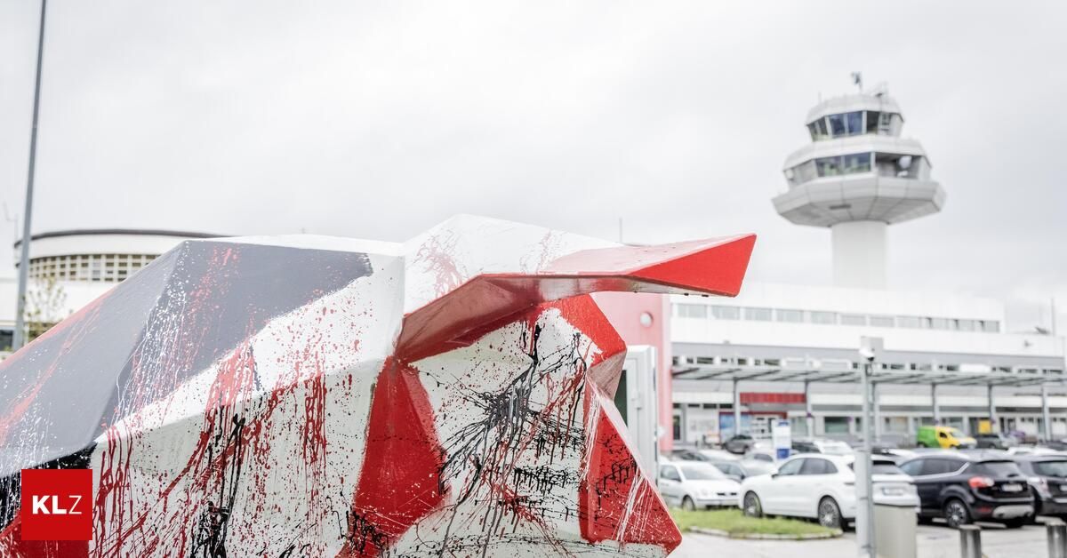 Ein geometrisches Kunstwerk mit roten und schwarzen Spritzern, gelegen an einem Flughafen mit geparkten Autos und einem Kontrollturm im Hintergrund.