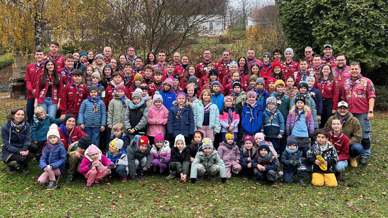 Eine große Gruppe von Kindern und Erwachsenen in Winterkleidung, möglicherweise Pfadfinder, posiert für ein Foto in einem Park mit Bäumen und einem Haus im Hintergrund.