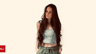 Eine Frau mit langen, welligen Haaren posiert für ein Foto in einem Jeans-Crop-Top und grünen Hosen. Sie hat einen ernsten Ausdruck und schaut geradeaus.