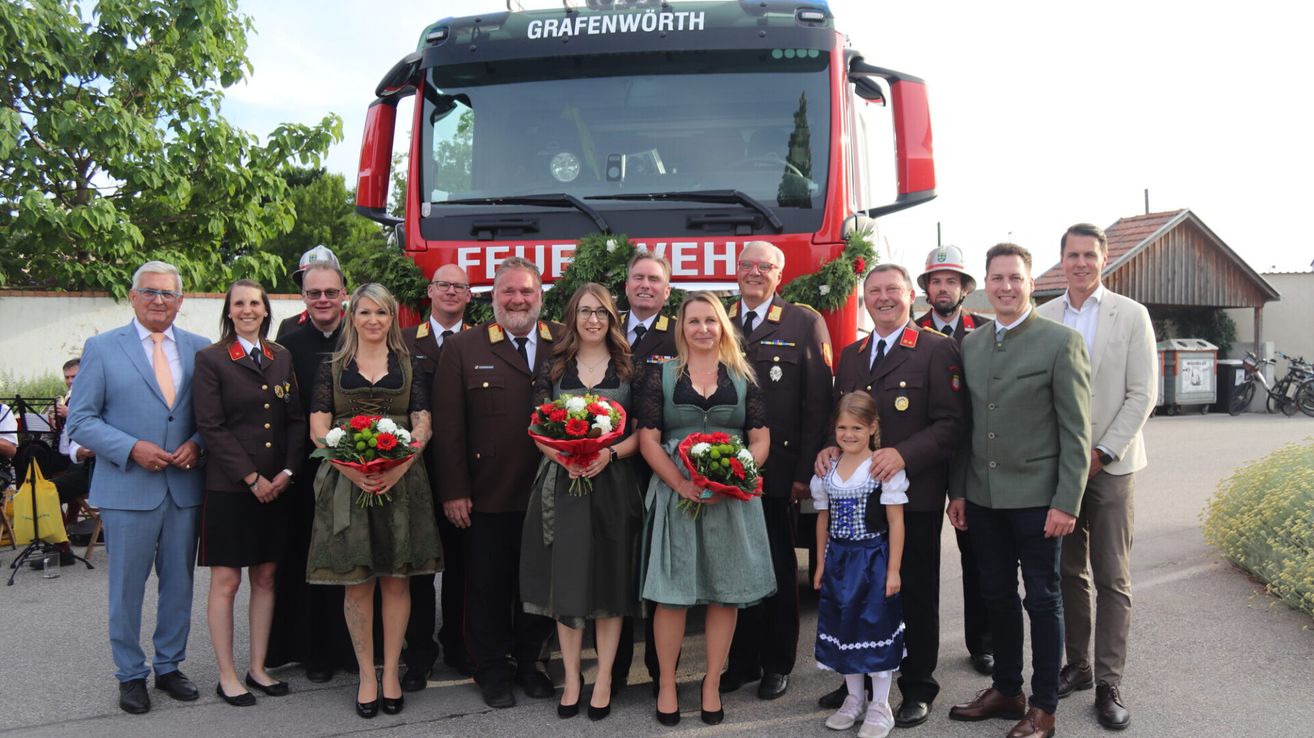 Eine Gruppe von Menschen in Uniformen steht vor einem Feuerwehrauto mit dem Schriftzug 'Grafenworth' und hält Blumen. Sie lächeln und scheinen für ein Foto zu posieren.