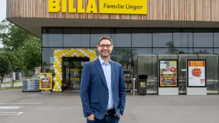 Ein Mann steht mit lächelndem Gesicht vor einem BILLA-Geschäft. Das Geschäft hat ein gelbes Schild mit dem Text BILLA Familie Unger. Mehrere Luftballons sind am Gebäude befestigt, und ein Mülleimer ist daneben platziert.