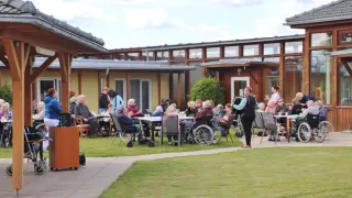 Eine Zusammenkunft älterer Menschen im Freien, die an Tischen unter einem bewölkten Himmel sitzen, mit einer Person in der Nähe, die einen Rucksack hält.