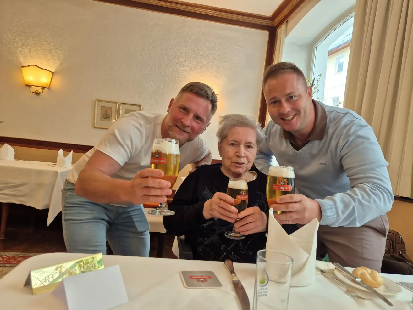 Drei Personen, zwei Männer und eine ältere Frau, lächeln und halten Biergläser in der Hand, wahrscheinlich zur Feier eines besonderen Anlasses. Sie sitzen an einem Tisch in einem Restaurant.