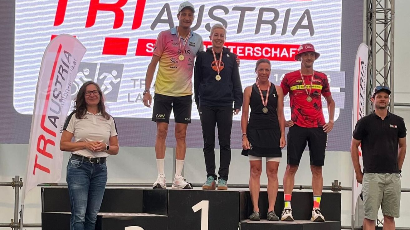 Fünf Personen stehen auf einem Podium und lächeln für die Kamera. Der Mann in der Mitte trägt eine Medaille um den Hals. Die Frau neben ihm hat eine Medaille um den Hals. Die Frau in Schwarz steht neben dem Mann in Rot.