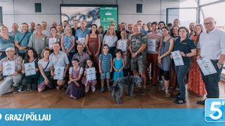 Eine große Gruppe von Menschen in Freizeitkleidung posiert für ein Foto. Sie halten Urkunden in den Händen, und einige tragen Abzeichen. Der Raum hat einen Holzboden und eine grüne Banner im Hintergrund.