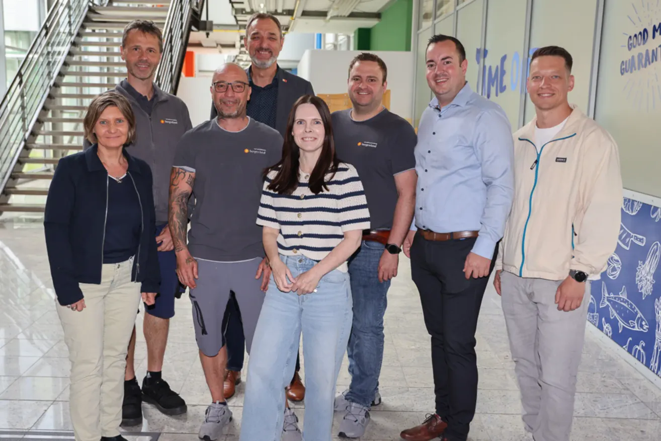 Eine Gruppe von Menschen, wahrscheinlich Kollegen, steht lächelnd zusammen in einem Flur. Die Frau in der Mitte trägt ein gestreiftes Shirt und Jeans. Die Männer sind lässig gekleidet. Der Hintergrund umfasst eine Treppe und eine Wand mit blauem und weißem Text.
