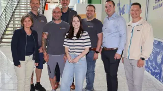 Eine Gruppe von Menschen, wahrscheinlich Kollegen, steht lächelnd zusammen in einem Flur. Die Frau in der Mitte trägt ein gestreiftes Shirt und Jeans. Die Männer sind lässig gekleidet. Der Hintergrund umfasst eine Treppe und eine Wand mit blauem und weißem Text.