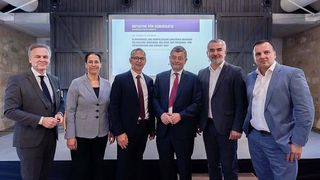 Sieben formell gekleidete Personen stehen lächelnd in einem Konferenzsaal vor einer Kamera. Dahinter zeigt eine Projektionsleinwand eine Präsentation mit dem Titel 'Initiative für Demokratie'.