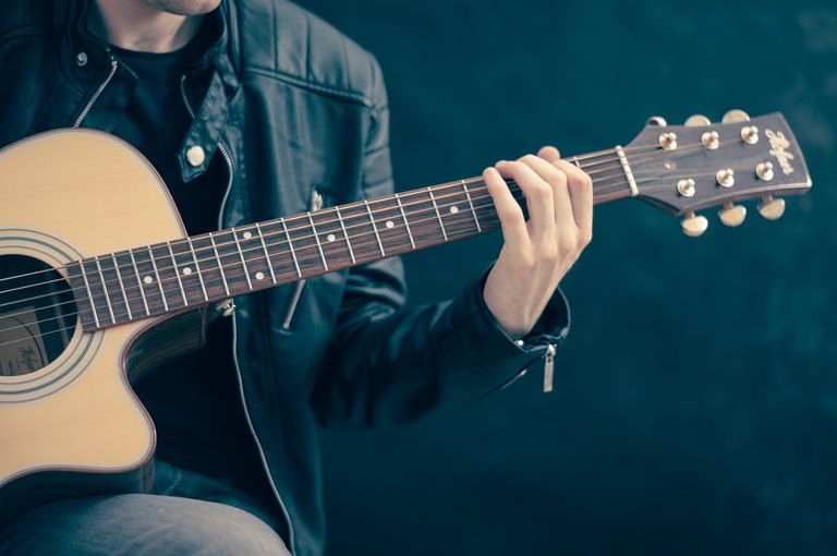 Eine Person in einer schwarzen Lederjacke spielt eine akustische Gitarre mit beiden Händen auf einem dunklen Hintergrund.