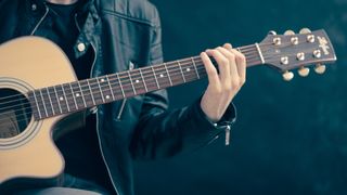 Eine Person in einer schwarzen Lederjacke spielt eine akustische Gitarre mit beiden Händen auf einem dunklen Hintergrund.