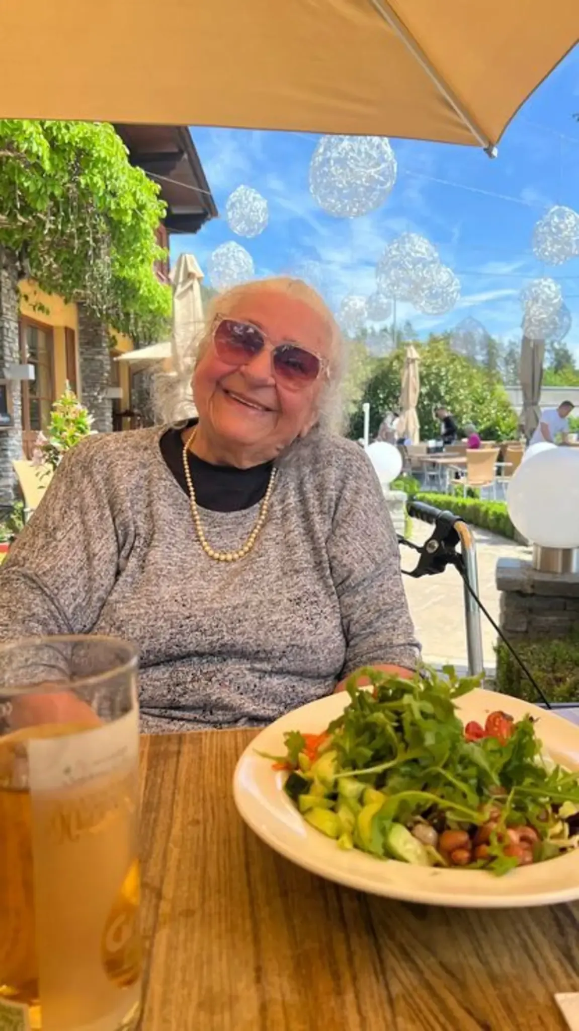 Eine ältere Frau in einem grauen Pullover und Sonnenbrille lächelt, während sie draußen in einem Restaurant sitzt. Ein Salat und ein Glas Bier stehen auf dem Tisch.